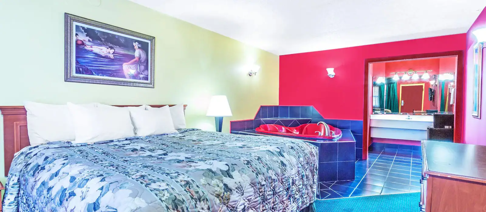 A guest room with a bed with a blue floral quilt, a red heart-shaped jacuzzi tub, and a bathroom with a vanity mirror