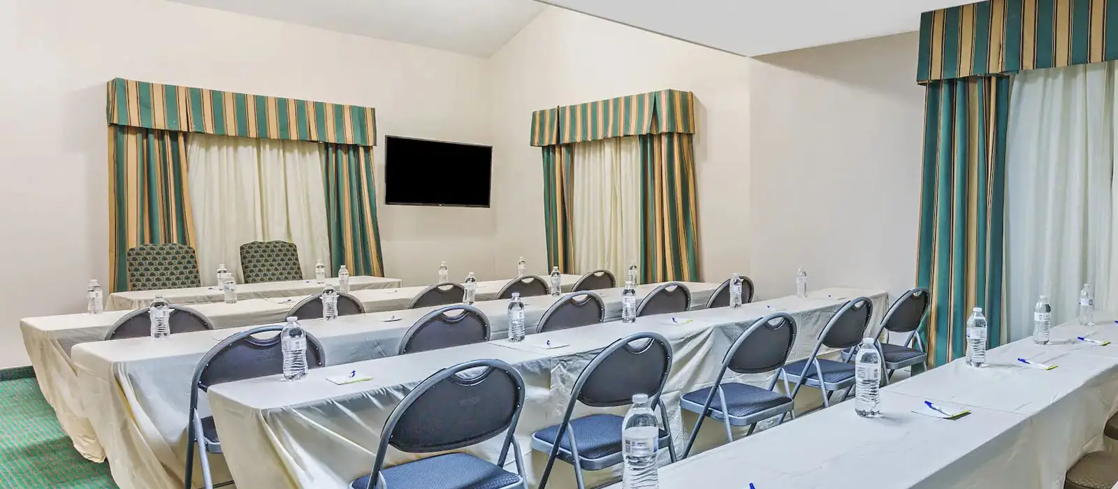 A conference room with several rows of tables covered in white cloths, chairs, and a wall-mounted television