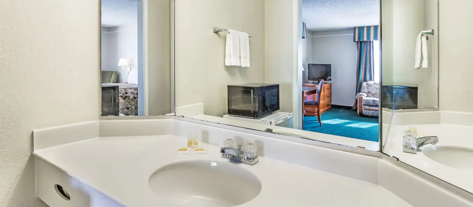 A bathroom vanity with a white sink and faucet, reflecting a microwave and a guest room with a bed and a TV