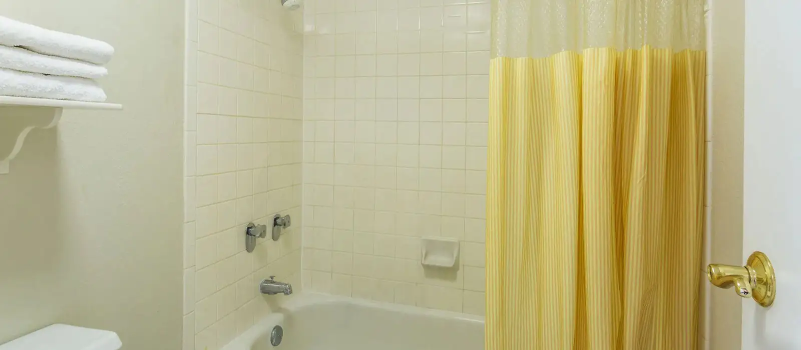 A white tiled bathroom with a bathtub, shower head, and a yellow and white striped shower curtain