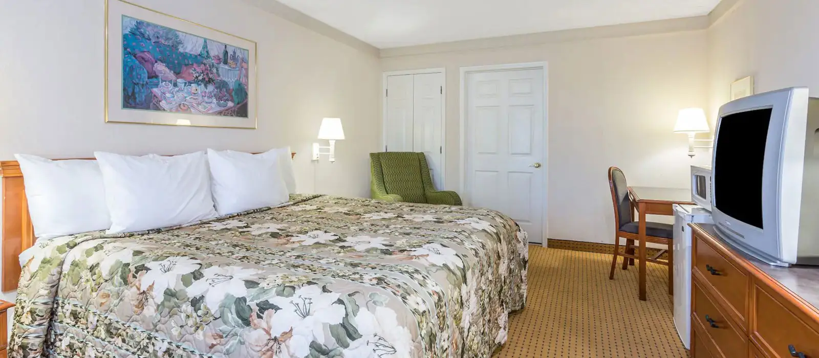A guest room with a bed in floral bedding, a green armchair, two white doors, a desk and chair, and a television on a dresser