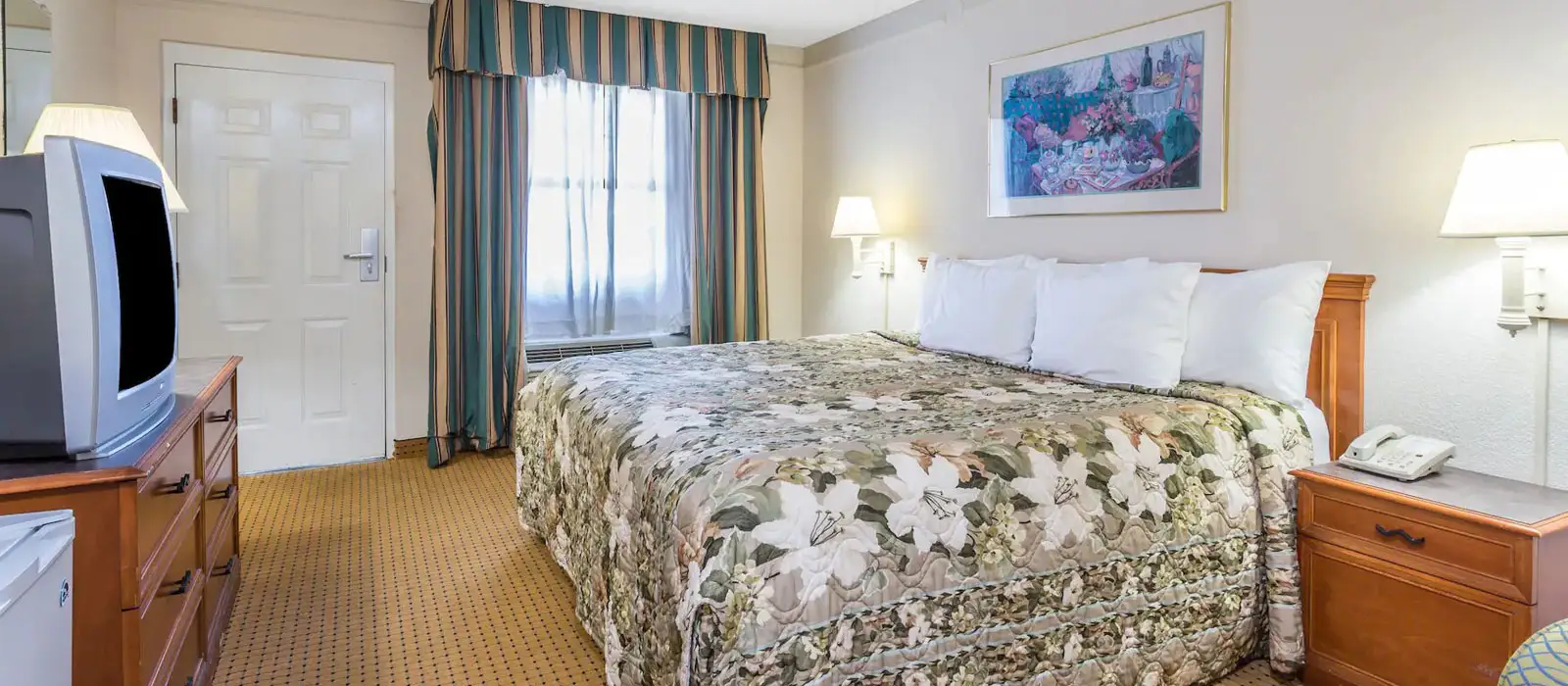 A guest room with a bed covered in a floral quilt, a window with striped curtains, a TV on a wooden dresser and a bedside table with a telephone