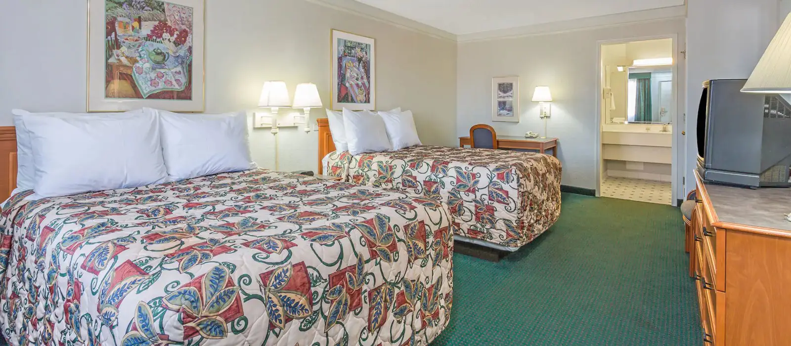 A hotel room with two beds with patterned bedspreads, a desk with a chair, and a bathroom visible through an open doorway