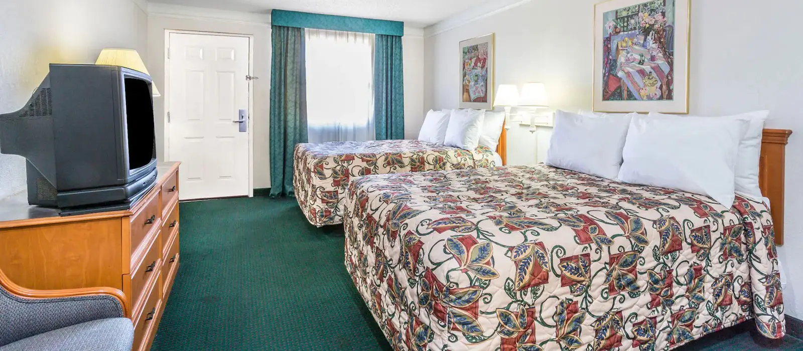 A guest room with two beds in patterned bedding, a television on a wooden dresser, and a window with teal curtains
