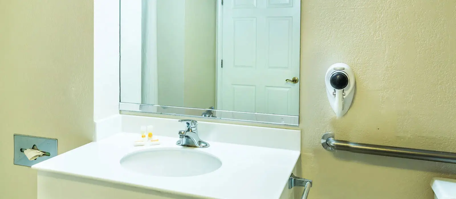 A bathroom vanity with a white sink, a mirror reflecting a white door, a wall-mounted tissue dispenser and a grab bar