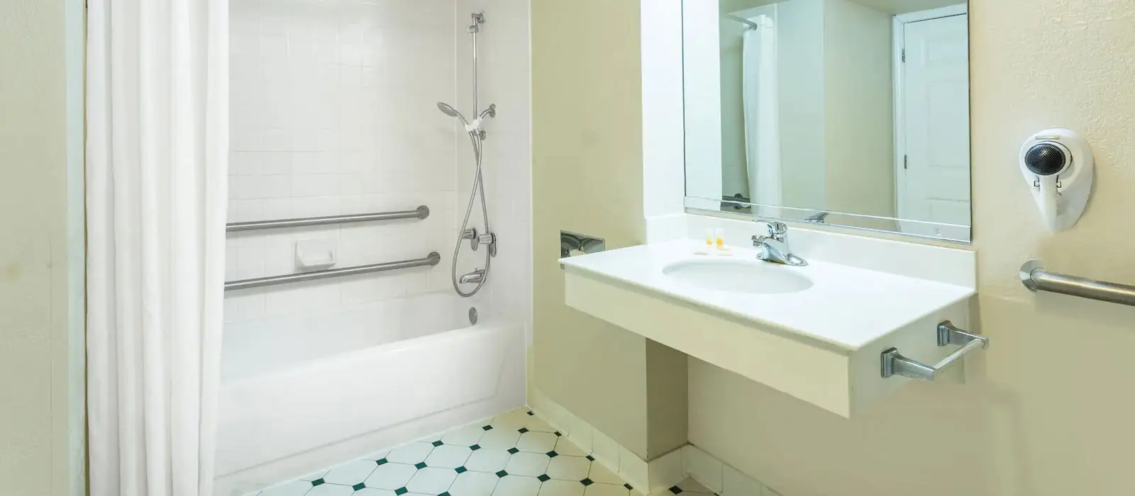 A bathroom with a white shower curtain, a bathtub with grab bars and a handheld shower, and a white vanity with a sink and mirror
