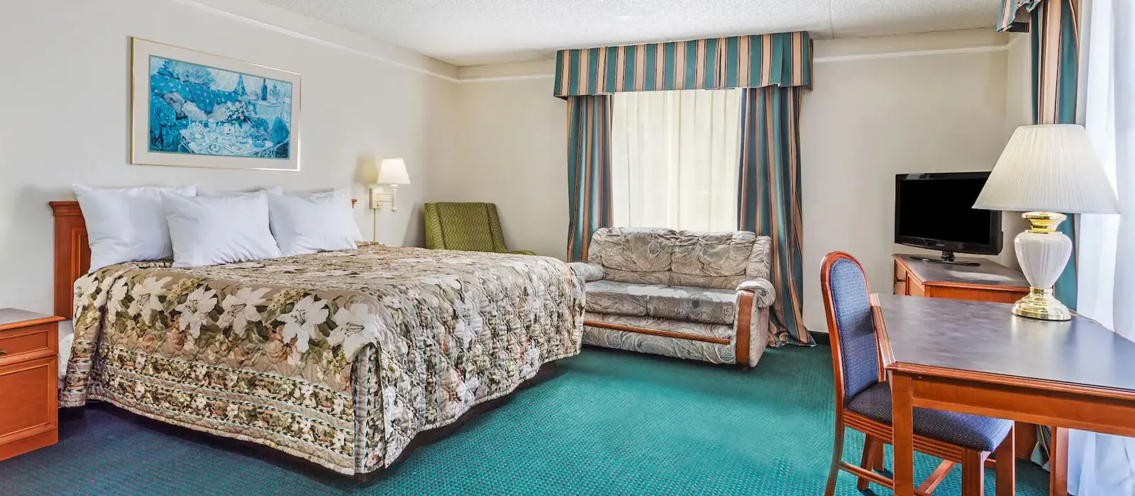A hotel room with a bed, a sofa, a green armchair, a desk with a chair, a television and a window with striped curtains