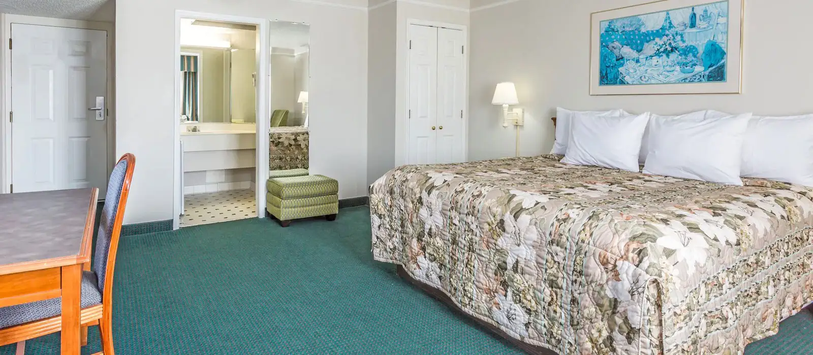 A guest room with a bed in floral bedding, a desk and chair, and an open doorway to a bathroom