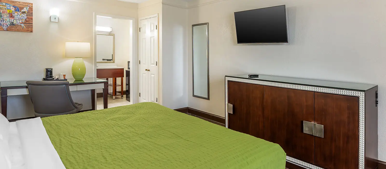 A guest room with a bed with a green bedspread, a desk with a coffee maker, a wall-mounted TV, and a dresser