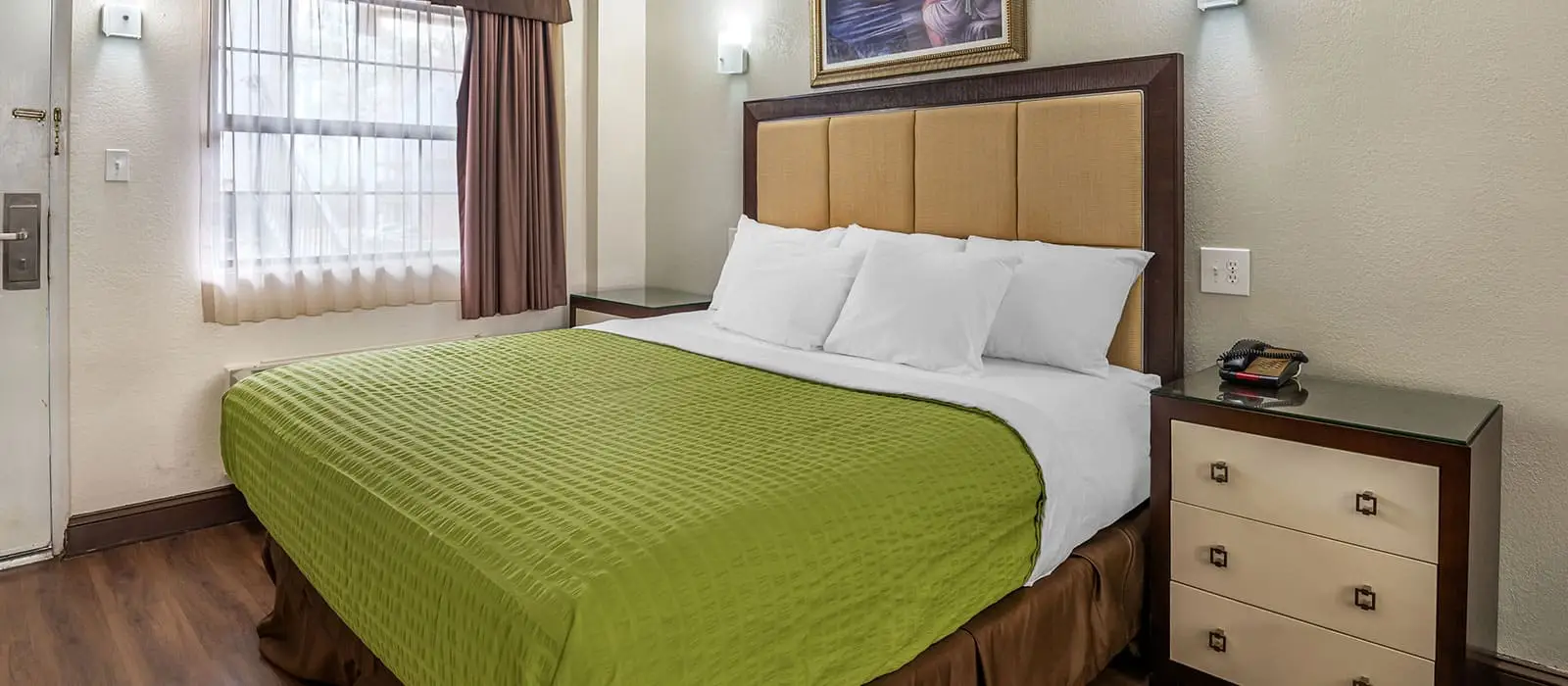 A hotel room with a bed covered in a green bedspread, a window with sheer and dark curtains, and a bedside table with a telephone