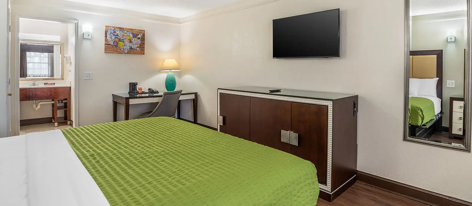 A guest room with a green bedspread, a desk with a lamp, a wall-mounted TV and a bathroom visible through an open doorway