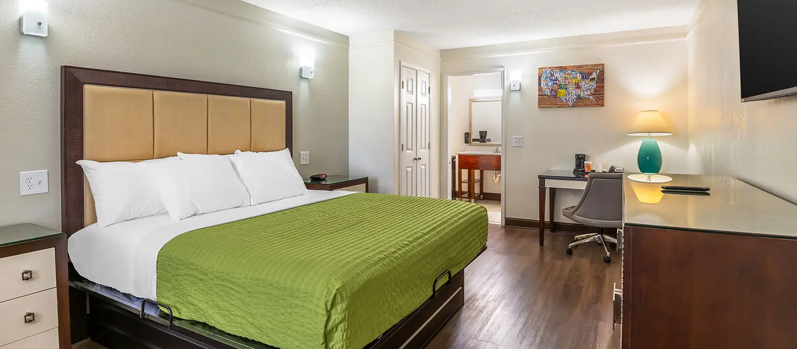 A guest room with a bed with a green bedspread, a desk with a chair, a TV and a vanity area with a sink