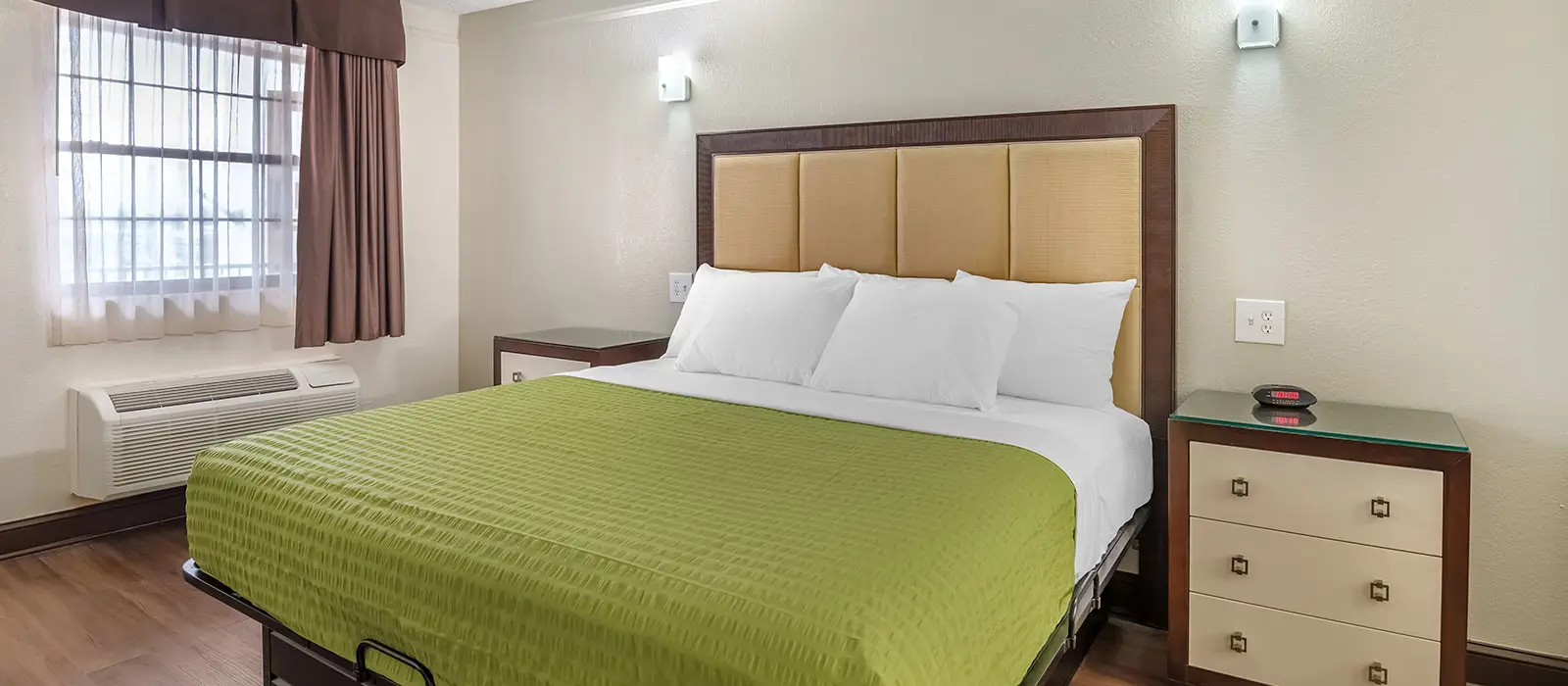 A guest room with a king bed in green and white bedding, two bedside tables, and a window with sheer and brown curtains