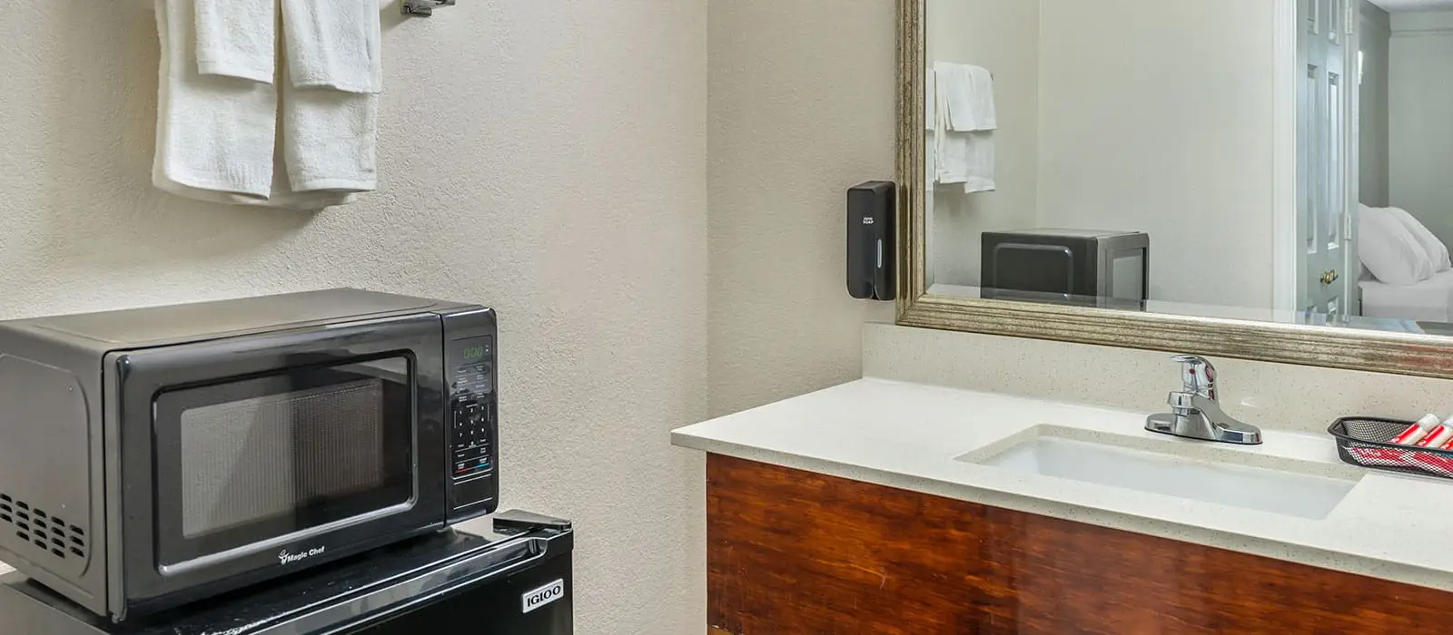 A microwave and mini-fridge sit below a towel rack, next to a vanity with a sink and mirror