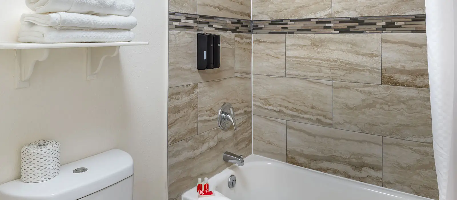 A bathroom with a white toilet, a white shelf with folded white towels, and a tiled shower with a white bathtub and two wall-mounted soap dispensers