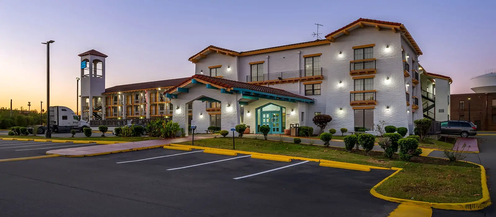 The hotel's white brick exterior with red tile roofs and teal accents, a parking lot, and a water tower in the distance