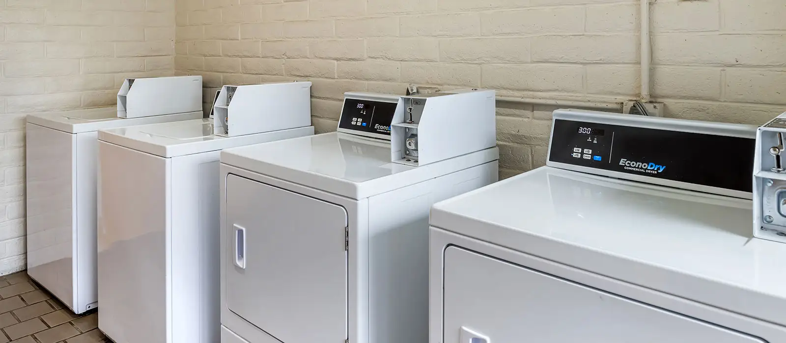 A row of white washing machines and dryers with coin slots and digital displays lines a light brick wall