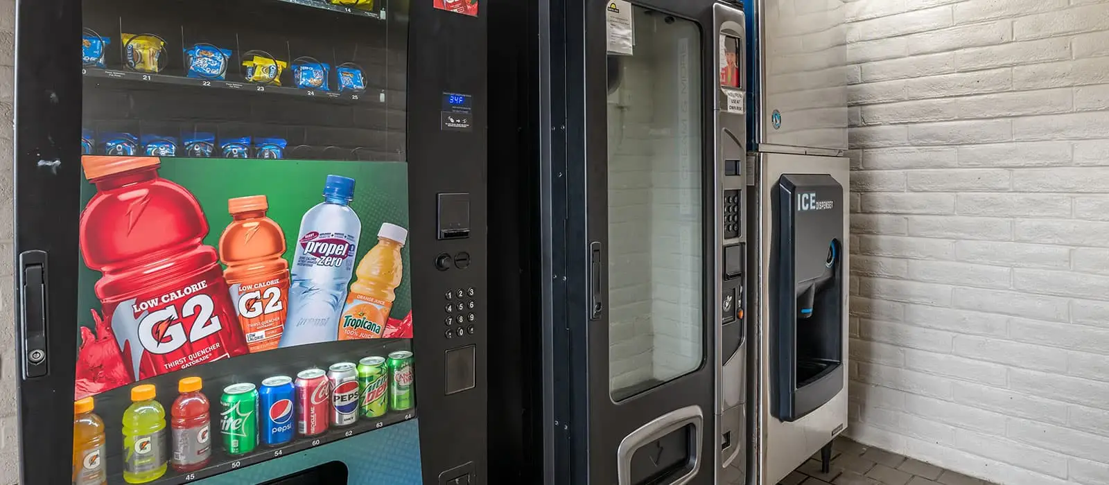 Two vending machines and an ice dispenser are against a white brick wall