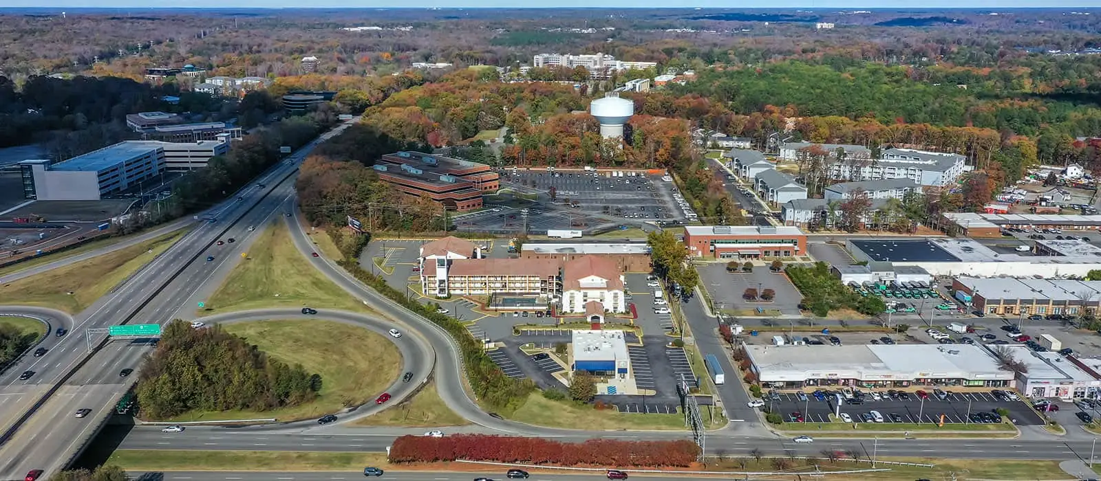 An aerial view of a highway interchange, buildings, and wooded areas under a blue sky