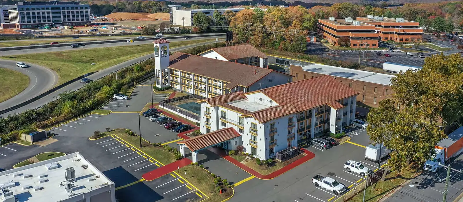 An aerial view of a hotel complex with multiple buildings, parking lots, a swimming pool and a highway in the background