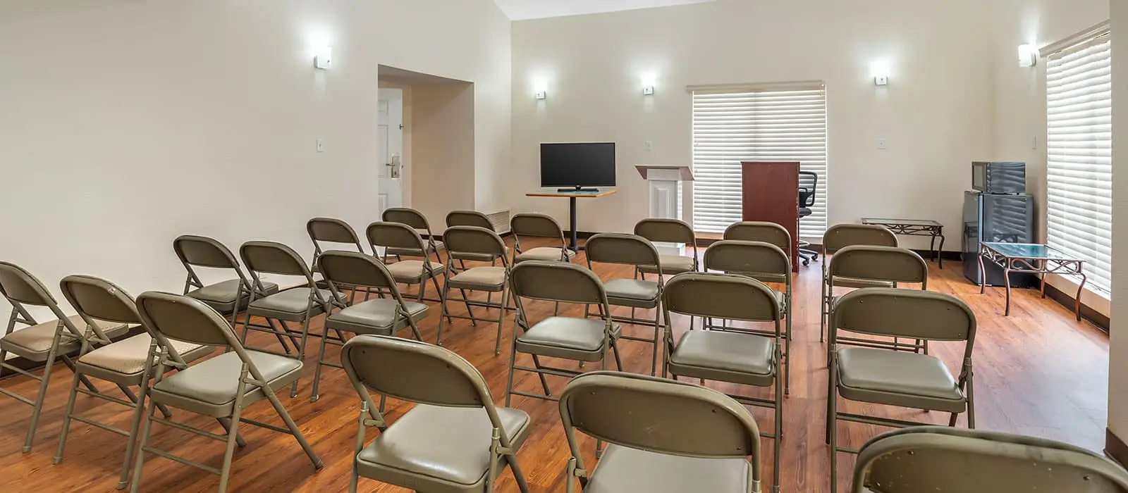 A meeting room with rows of grey folding chairs facing a television on a stand, a white podium and a wooden lectern