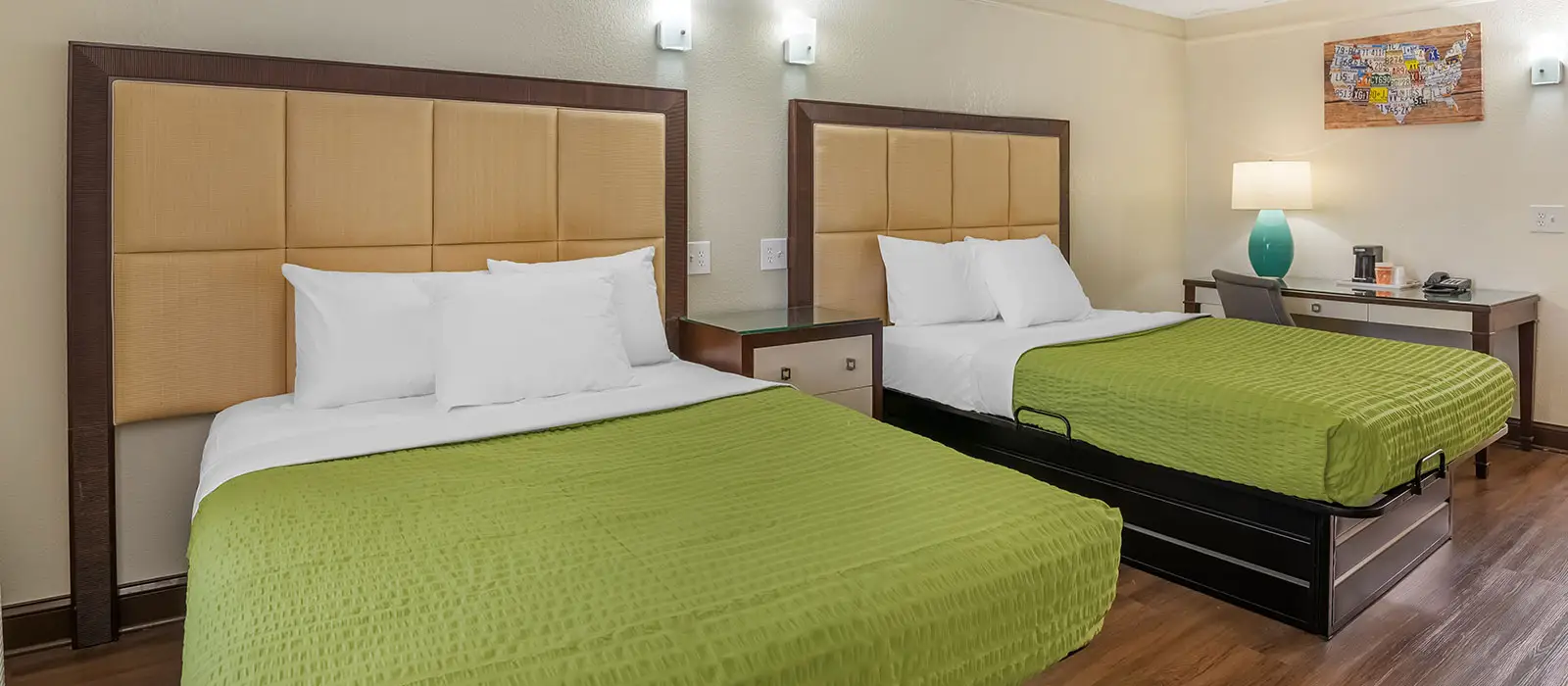 A guest room with two beds with green bedspreads, a bedside table and a desk with a lamp and coffee maker