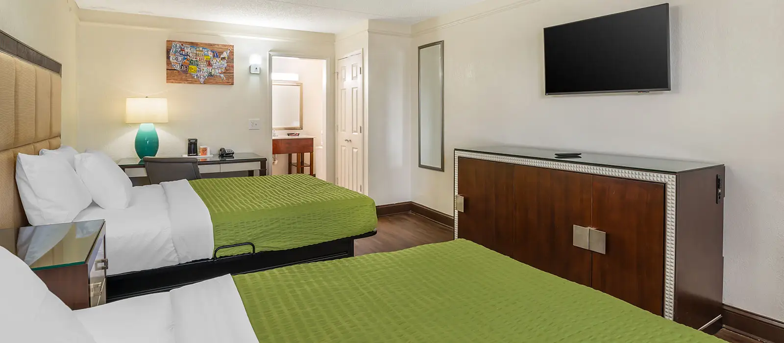 A guest room with two beds with green bedspreads, a desk with a lamp, a TV and a dresser
