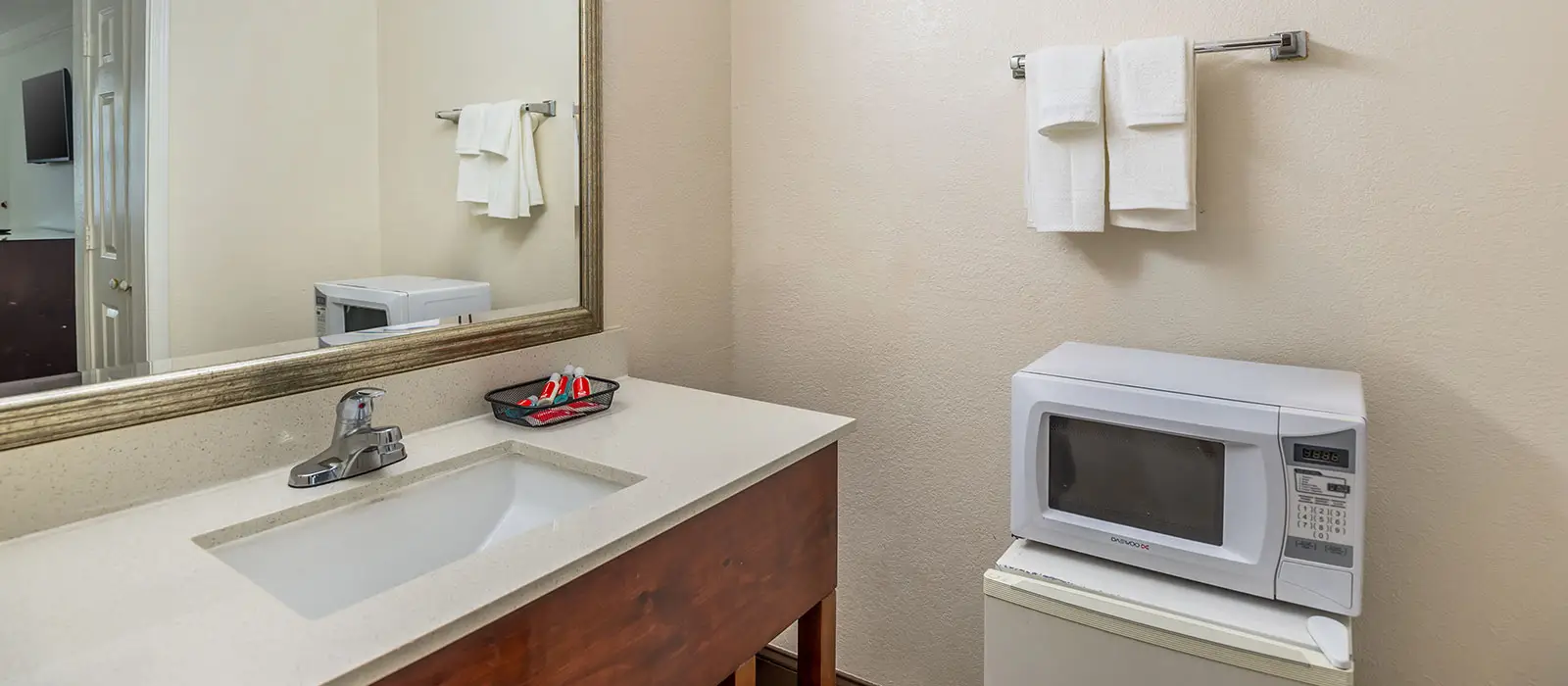 A bathroom vanity with a sink and faucet, a mirror reflecting a microwave, and a microwave and mini-fridge to the right