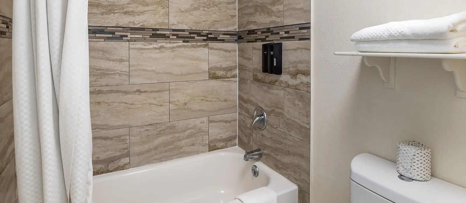 A bathroom with a white bathtub, beige tiled walls, a shower curtain and a shelf with white towels