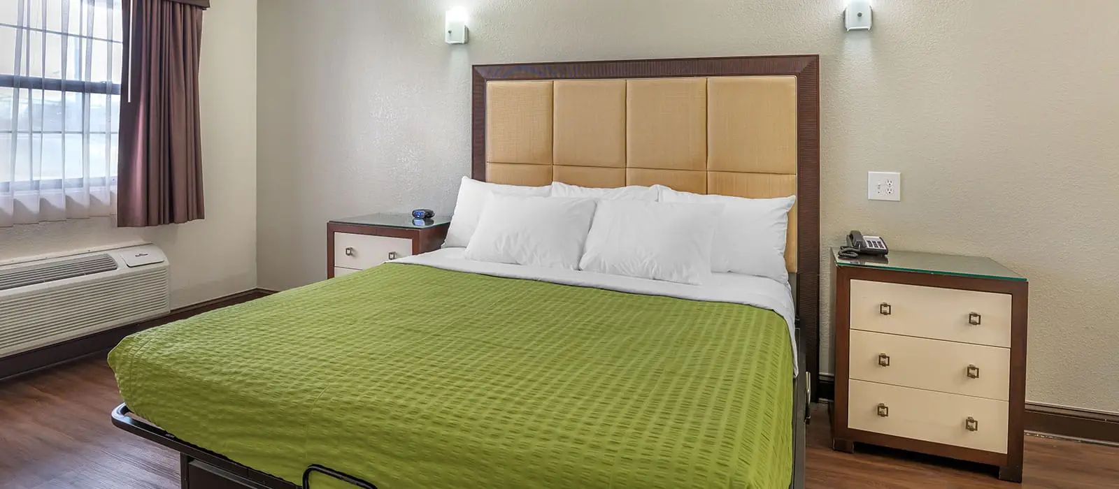 A hotel room with a bed covered in a green blanket, two bedside tables, and a window with sheer and brown curtains