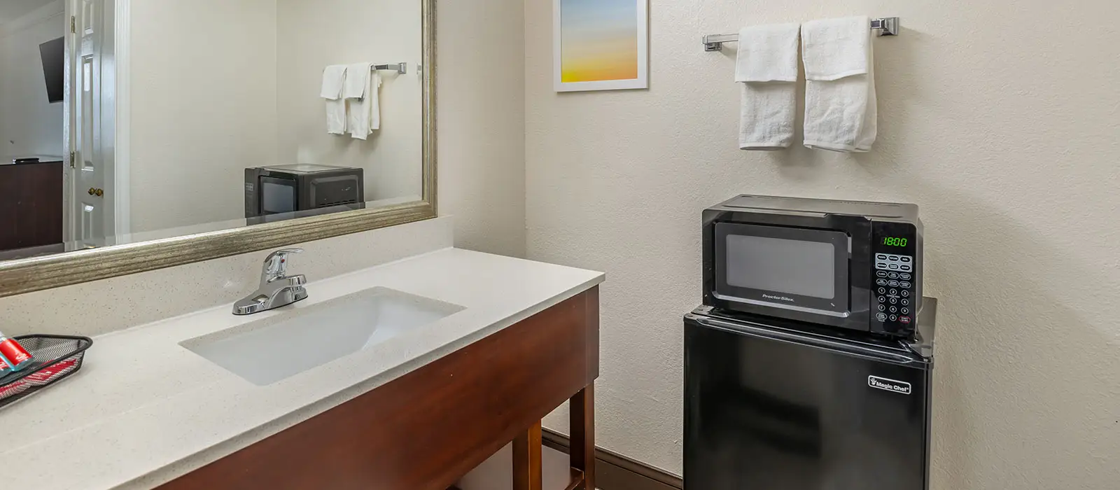 A bathroom vanity with a sink and faucet, a mirror, and a microwave and mini-fridge unit
