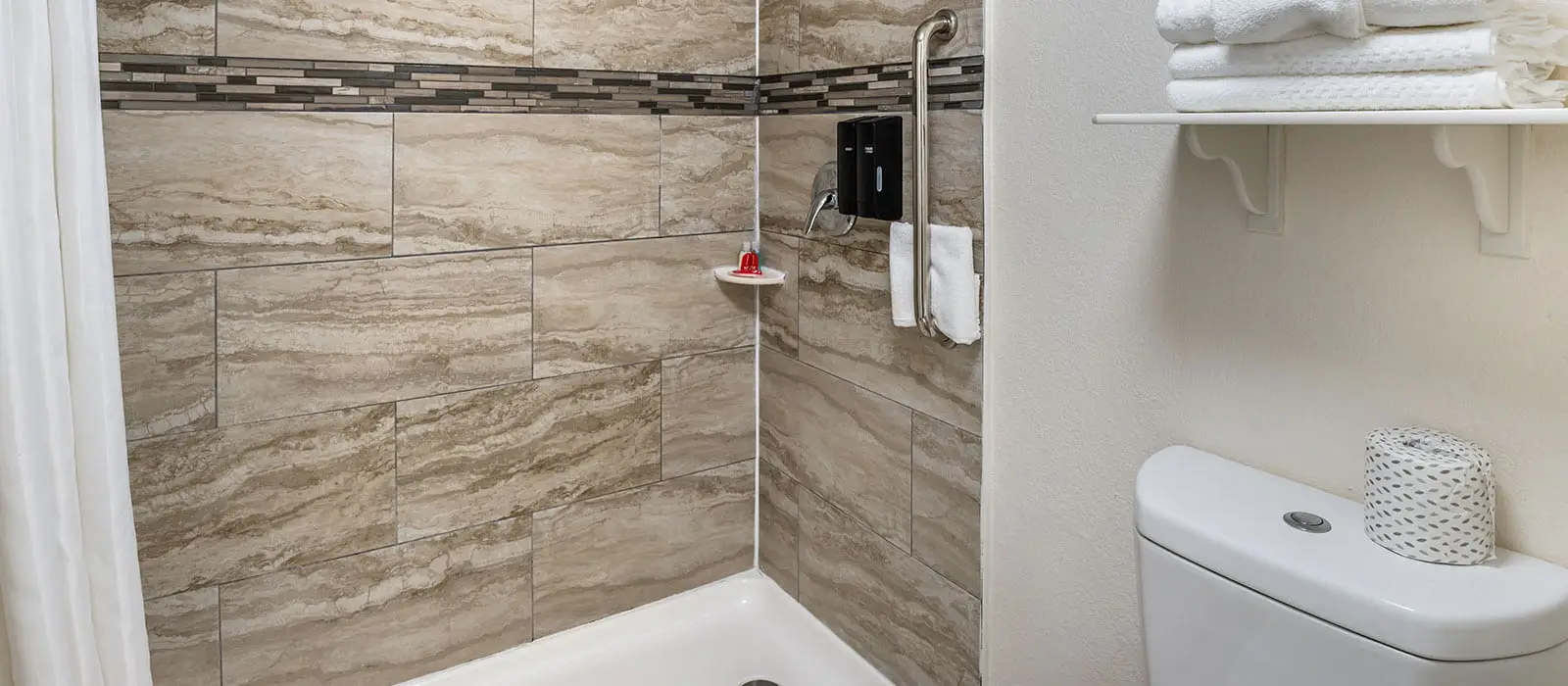 A bathroom with a shower tiled in beige and brown, a grab bar, two soap dispensers and a toilet with a roll of toilet paper