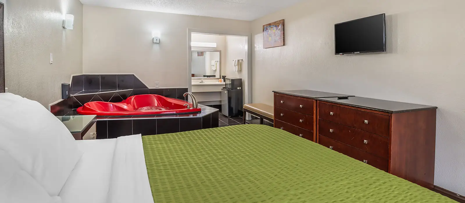 A guest room with a red corner hot tub, a bed with a green bedspread, a dresser, a wall-mounted TV and a bathroom entrance