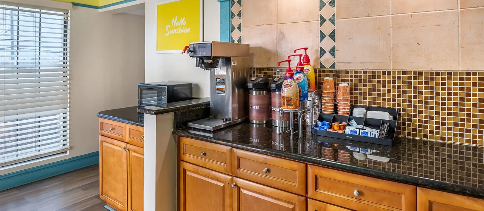 A coffee station with a microwave, a coffee maker, coffee supplies and paper cups on a dark granite counter