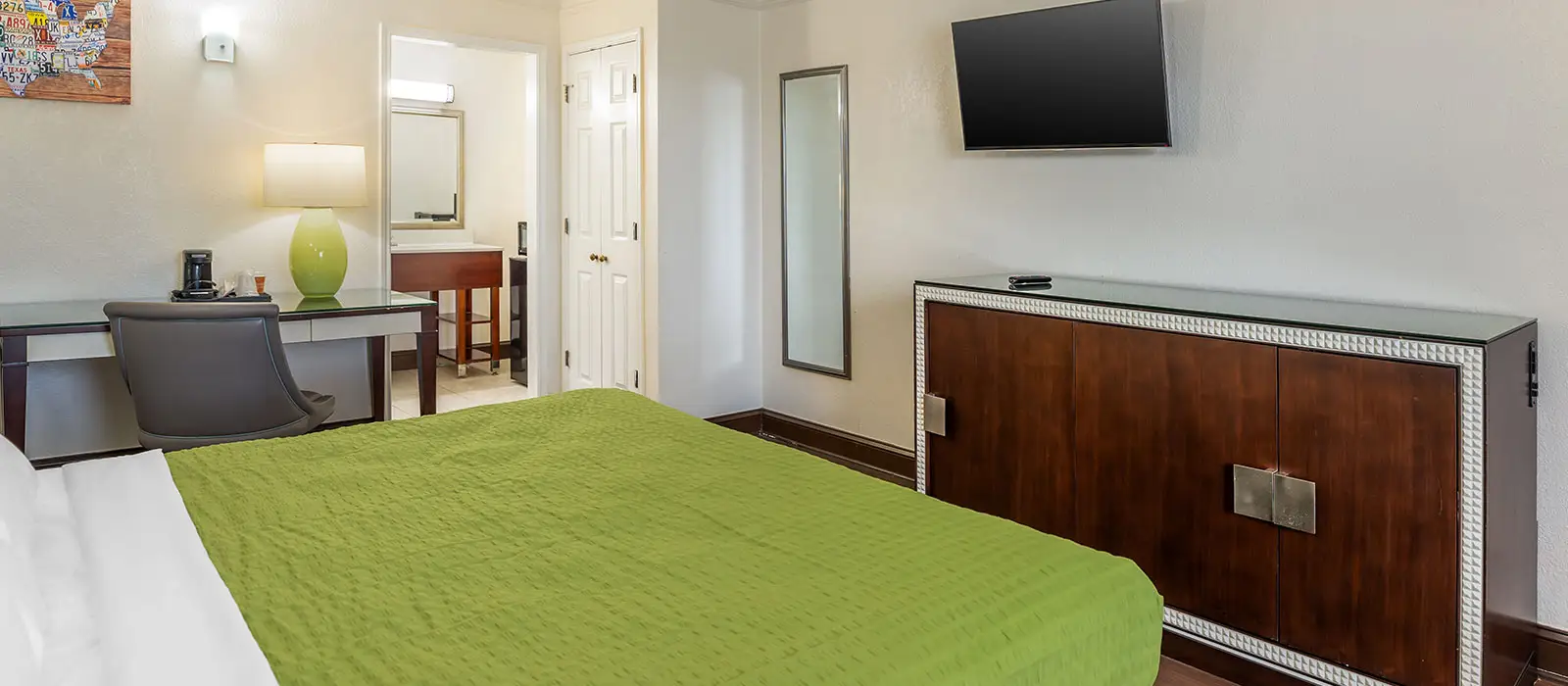 A hotel room with a green bedspread, a desk with a chair, a wall-mounted television and a partial view of a bathroom