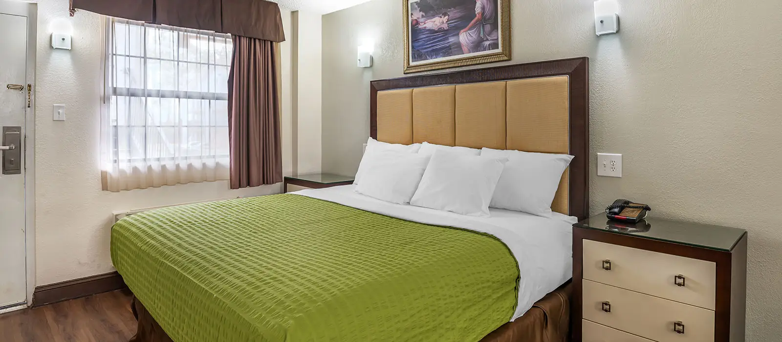 A guest room with a bed covered in a green blanket and white pillows, a window with sheer and dark brown curtains, and a bedside table with a phone