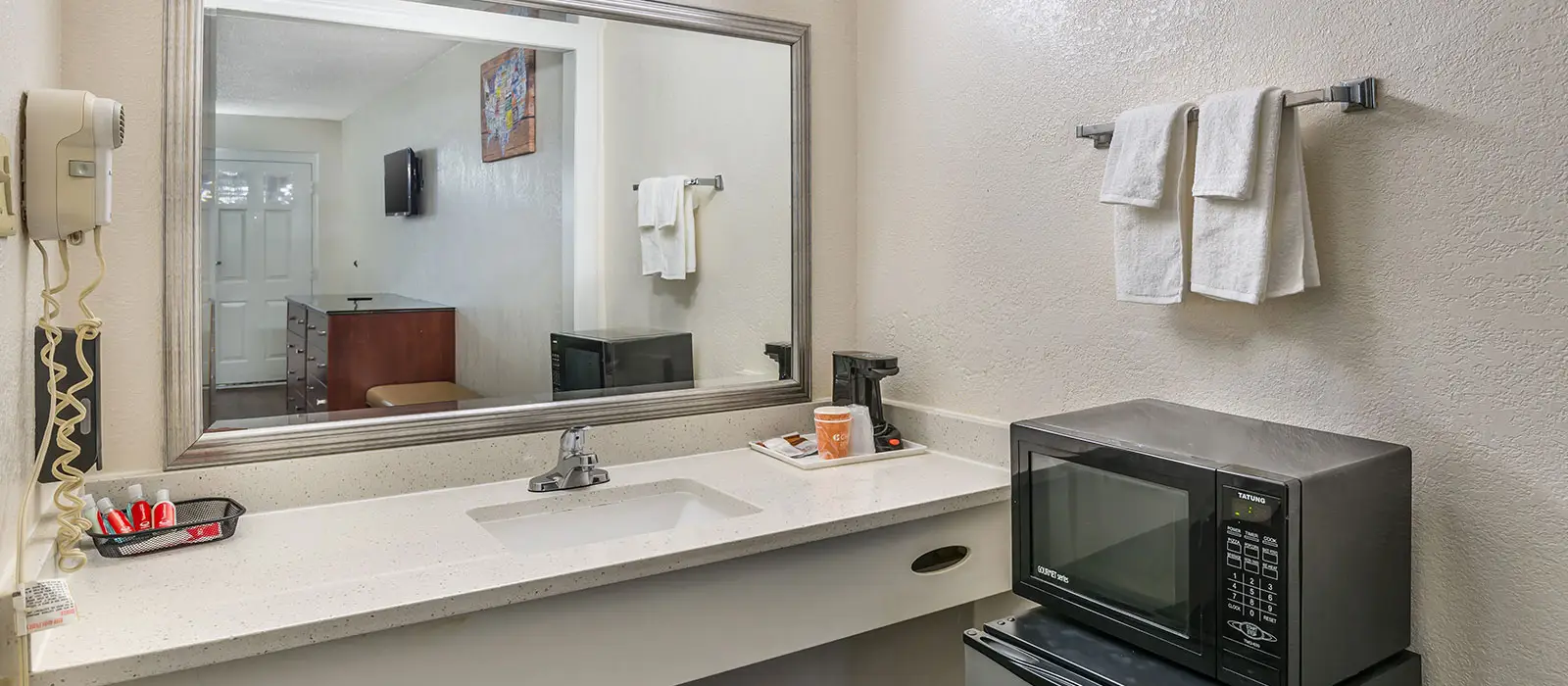 A bathroom vanity with a sink, a hairdryer, a coffee maker, a microwave and a towel rack with two white towels