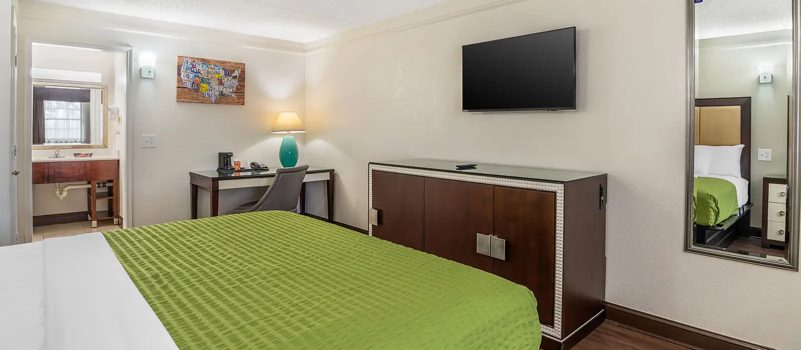 A hotel room with a green bedspread, a desk with a lamp, a wall-mounted TV above a dresser and a bathroom visible through an open doorway