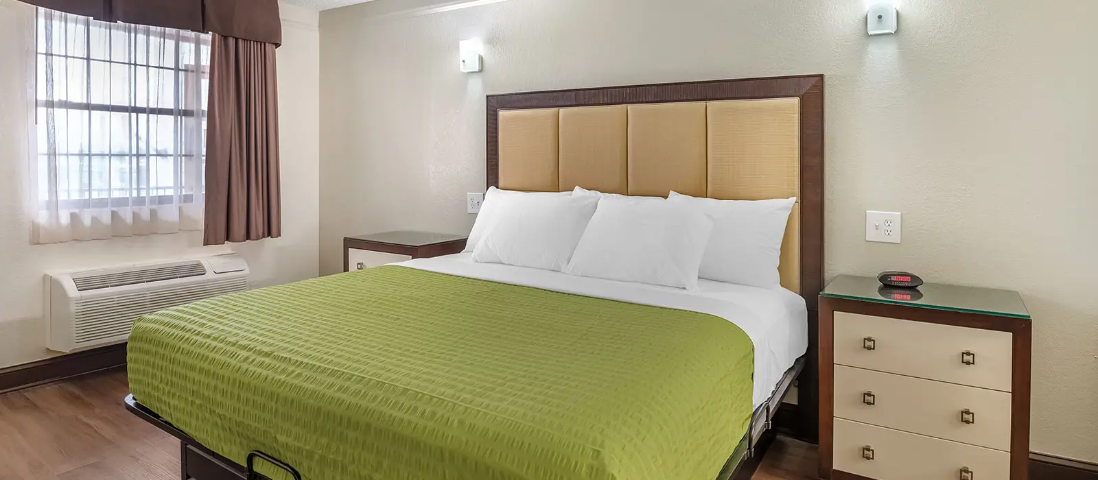 A guest room with a bed with a green bedspread, a window with sheer and brown curtains, and two bedside tables