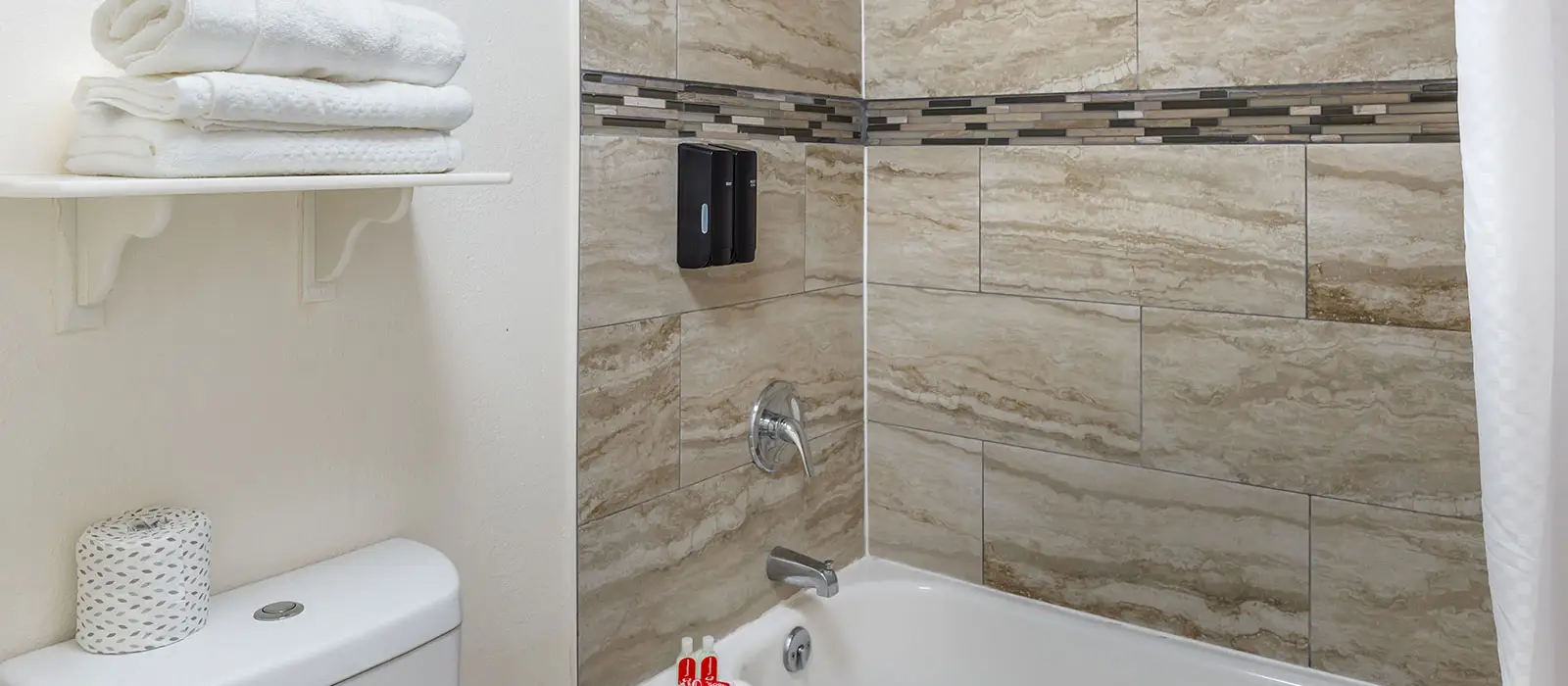 A bathroom with a white toilet, a shelf with folded white towels, and a bathtub with tiled walls