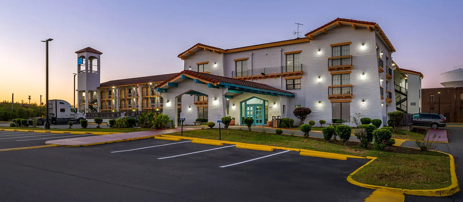The hotel exterior at dusk, featuring a white building with a red tile roof, teal accents and a parking lot in the foreground
