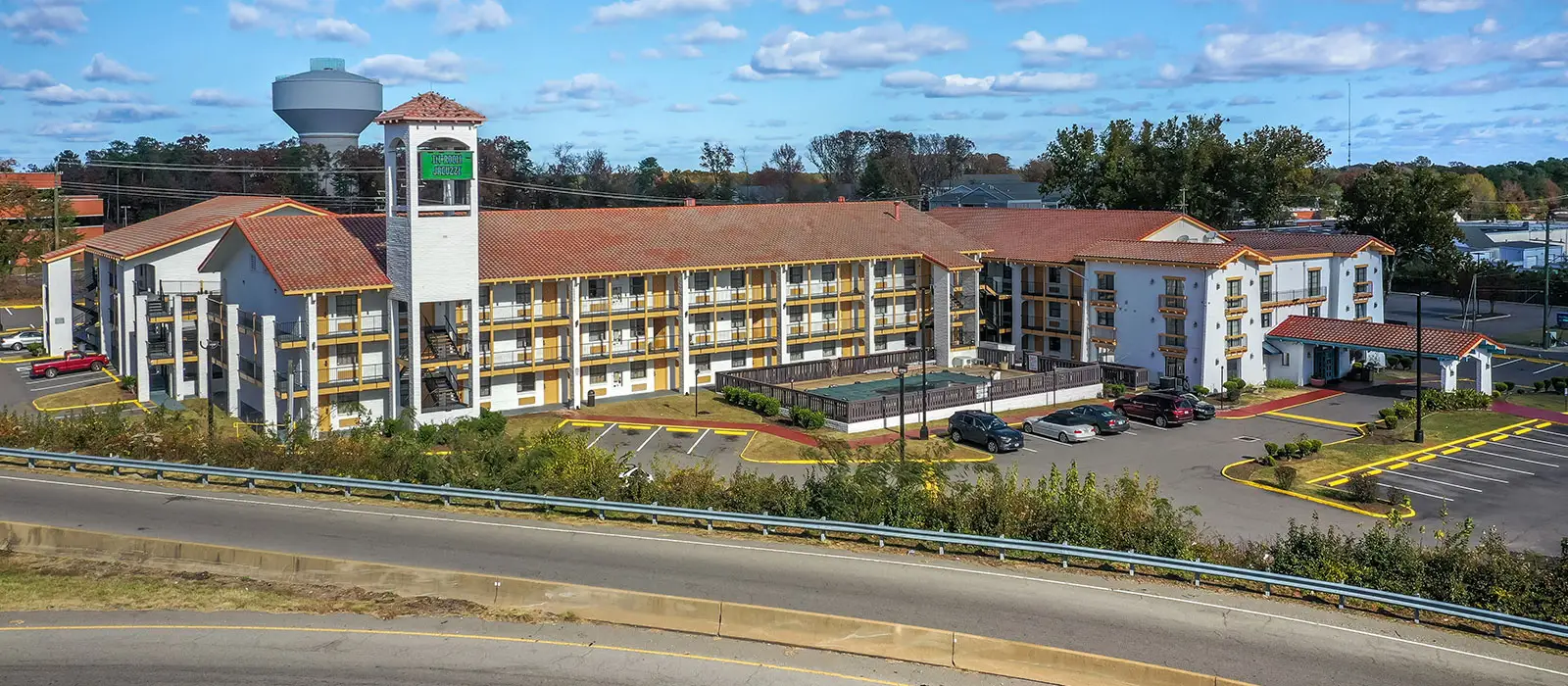 An aerial view of a multi-storey hotel with a red tiled roof, a swimming pool, and a parking lot, next to a road