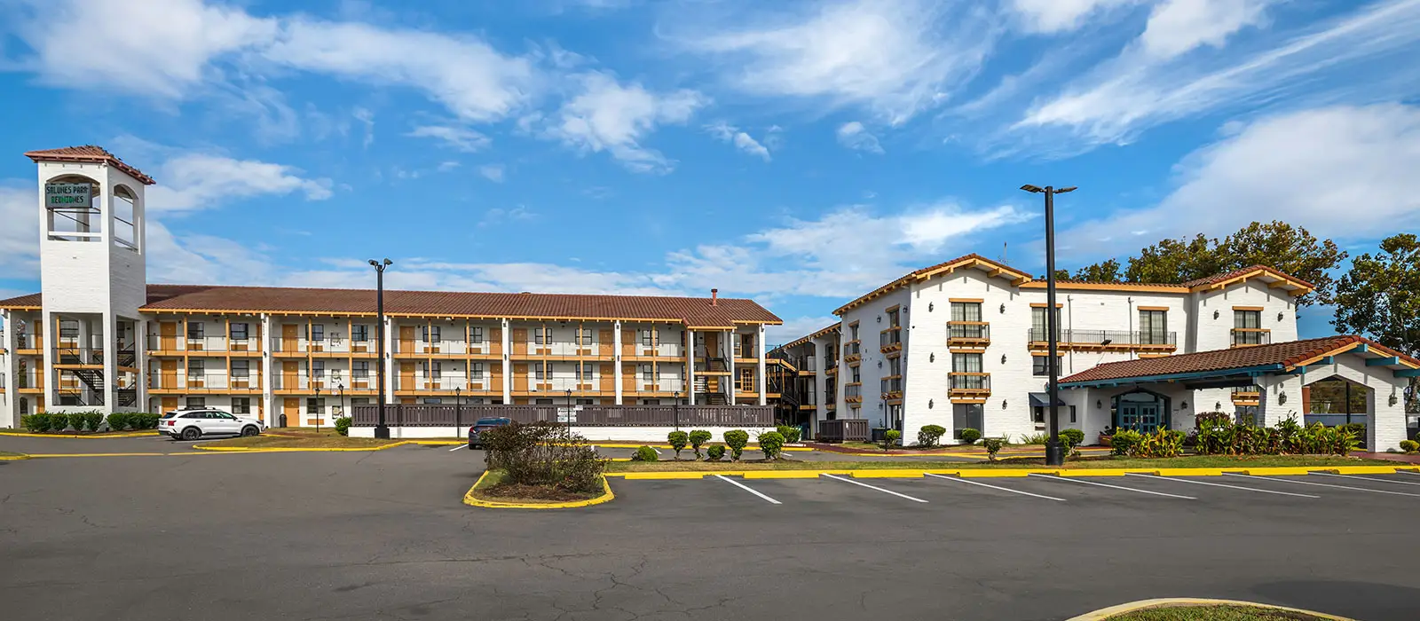 Two white two-storey buildings with brown tiled roofs and balconies, separated by a parking area, under a blue sky