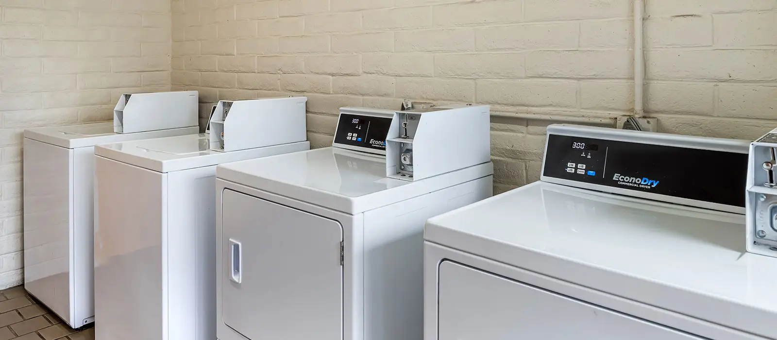 A row of white washing machines and dryers with coin slots in a laundry room with light brick walls