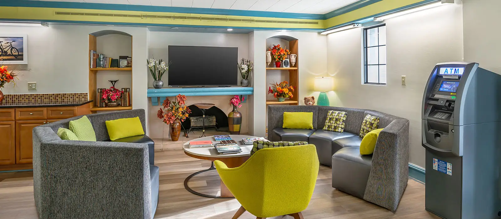 A lounge area with grey sofas, a yellow armchair, a coffee table, a television, built-in shelves and an ATM