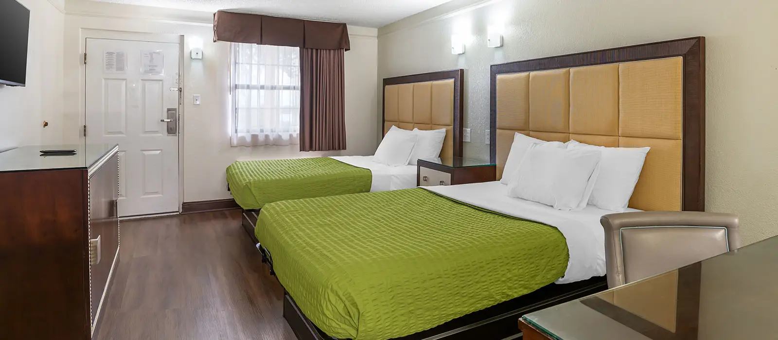A guest room with two beds in green bedding, a window with sheer and dark curtains, a TV and a desk with a chair