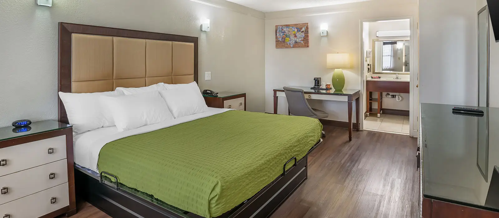 A guest room with a bed in green bedding, a desk with a chair, and a partial view of a bathroom vanity