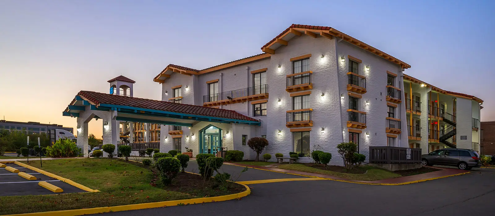 The hotel exterior at dusk, showing a white brick building with red tile roofs, balconies, and a covered entrance