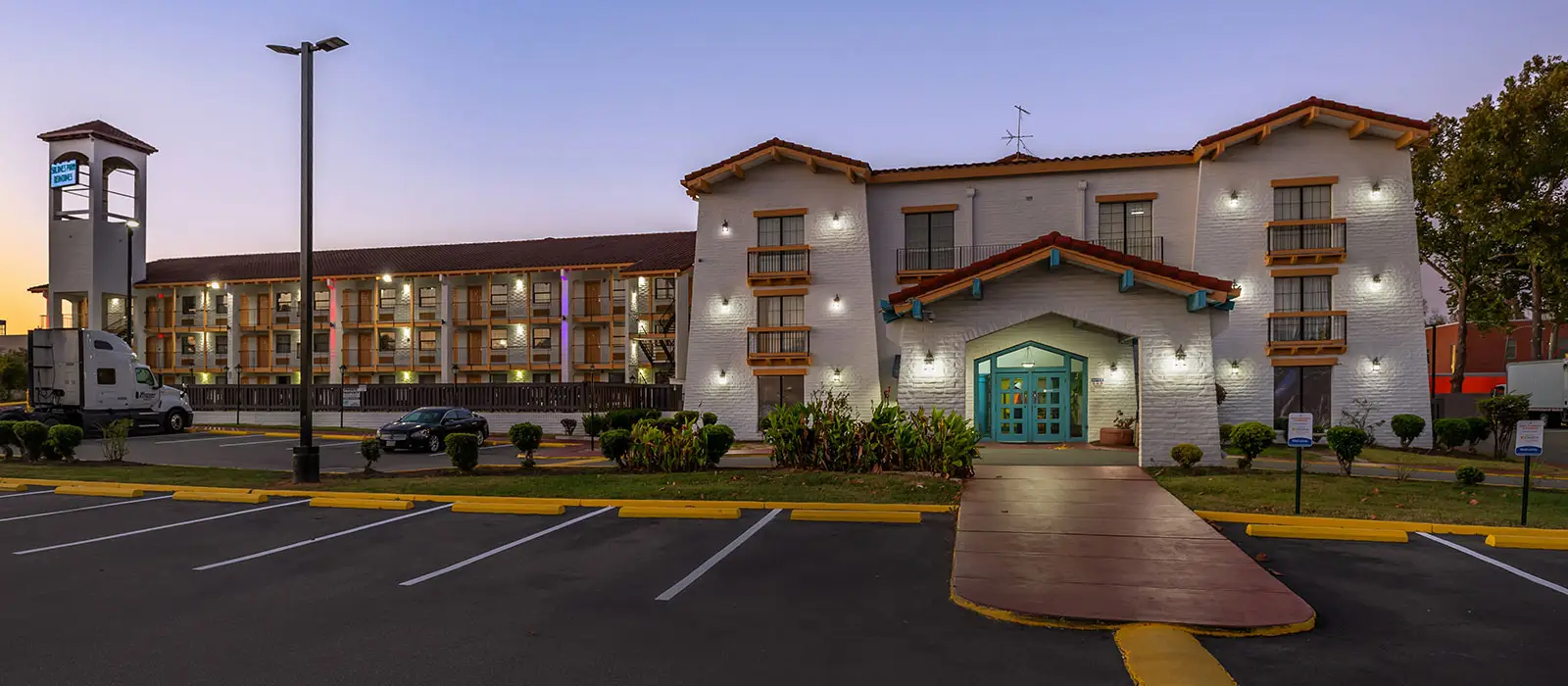 The hotel exterior at dusk, with a white building featuring a red-tiled roof, balconies, and a turquoise entrance, next to a parking lot with a parked truck and car