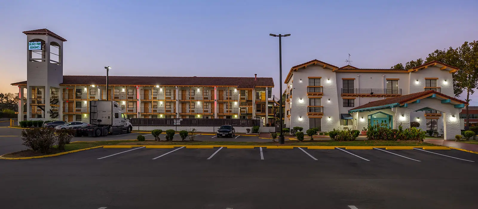 Two white two-storey buildings with red tiled roofs and a parking lot in the foreground, under a twilight sky
